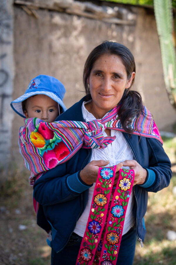 Dame bærer barn på ryggen og viser håndarbejde frem