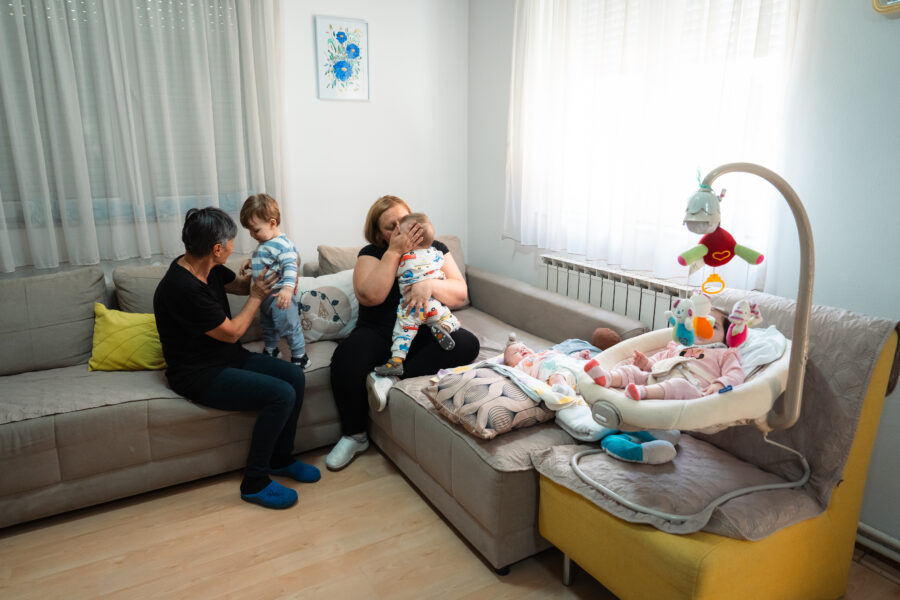 To kvinder sidder på en sofa og holder hvert sit lille barn. På sofaen ligger en baby på et turkis babytæppe, en anden baby ligger i en babystol placeret på sofaen. 