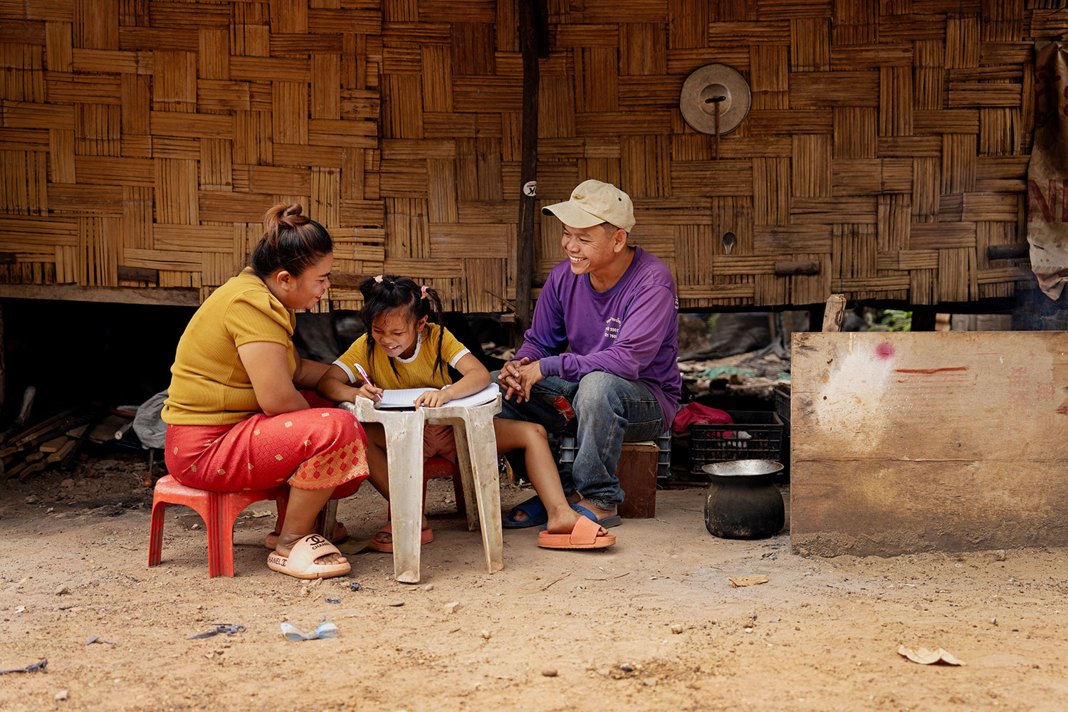 Billedet viser en familie i Laos, som får støtte af SOS Børnebyerne. Moren, faren og deres datter på seks år sidder foran familiens hus og laver lektier ved et lille, hvidt plastikbord. De griner og hygger sig sammen.